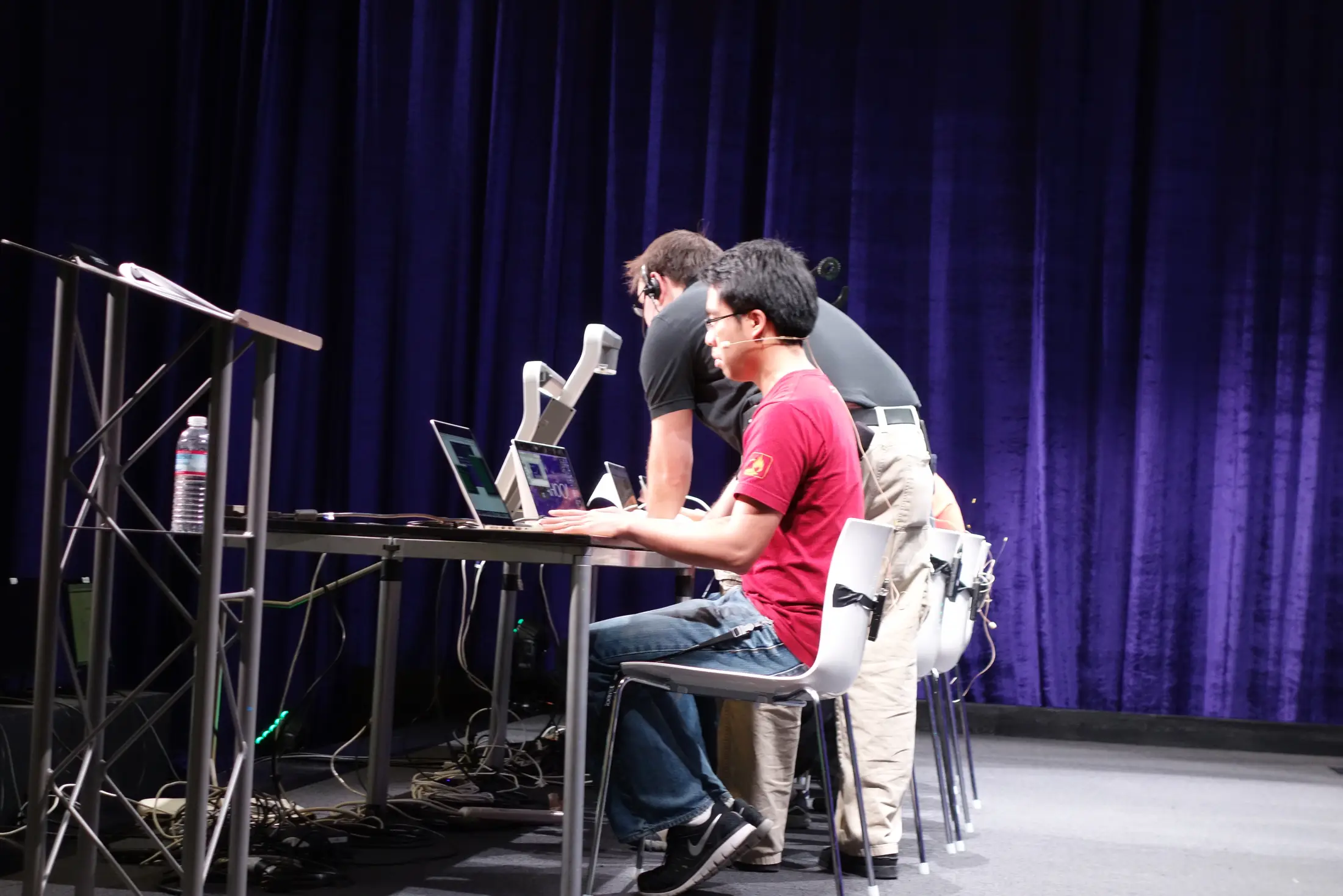 Hackers building on stage at a hackathon, against a deep blue curtain